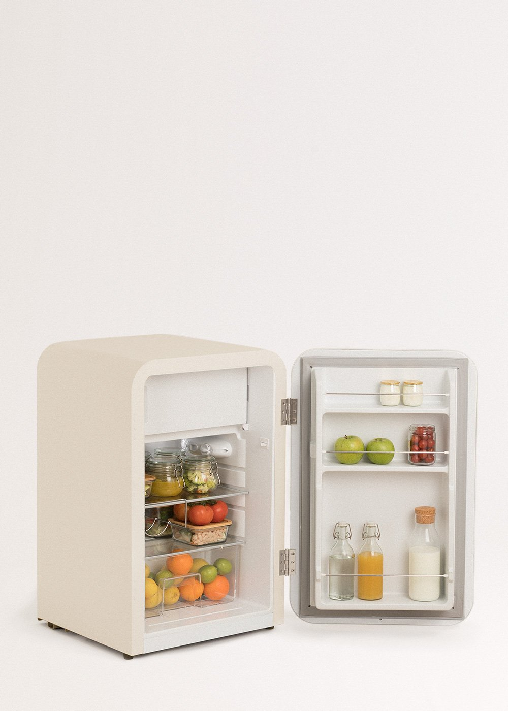 Un pequeño refrigerador crema abierto lleno de frutas, verduras y frascos. El fondo es blanco.