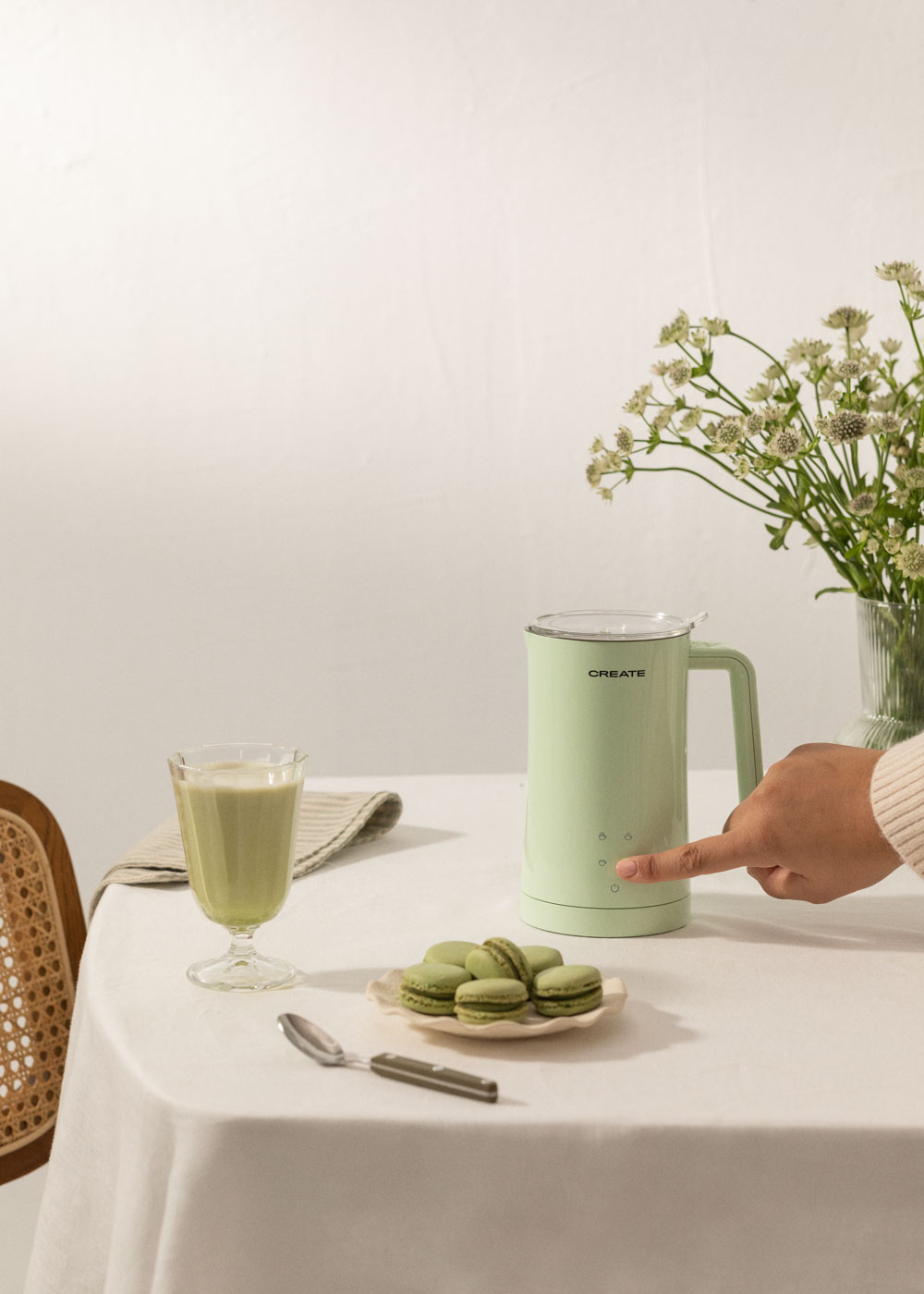 Un electrodoméstico verde menta está sobre una mesa. Una mano presiona un botón. Macarons, una bebida y flores también están en la mesa.