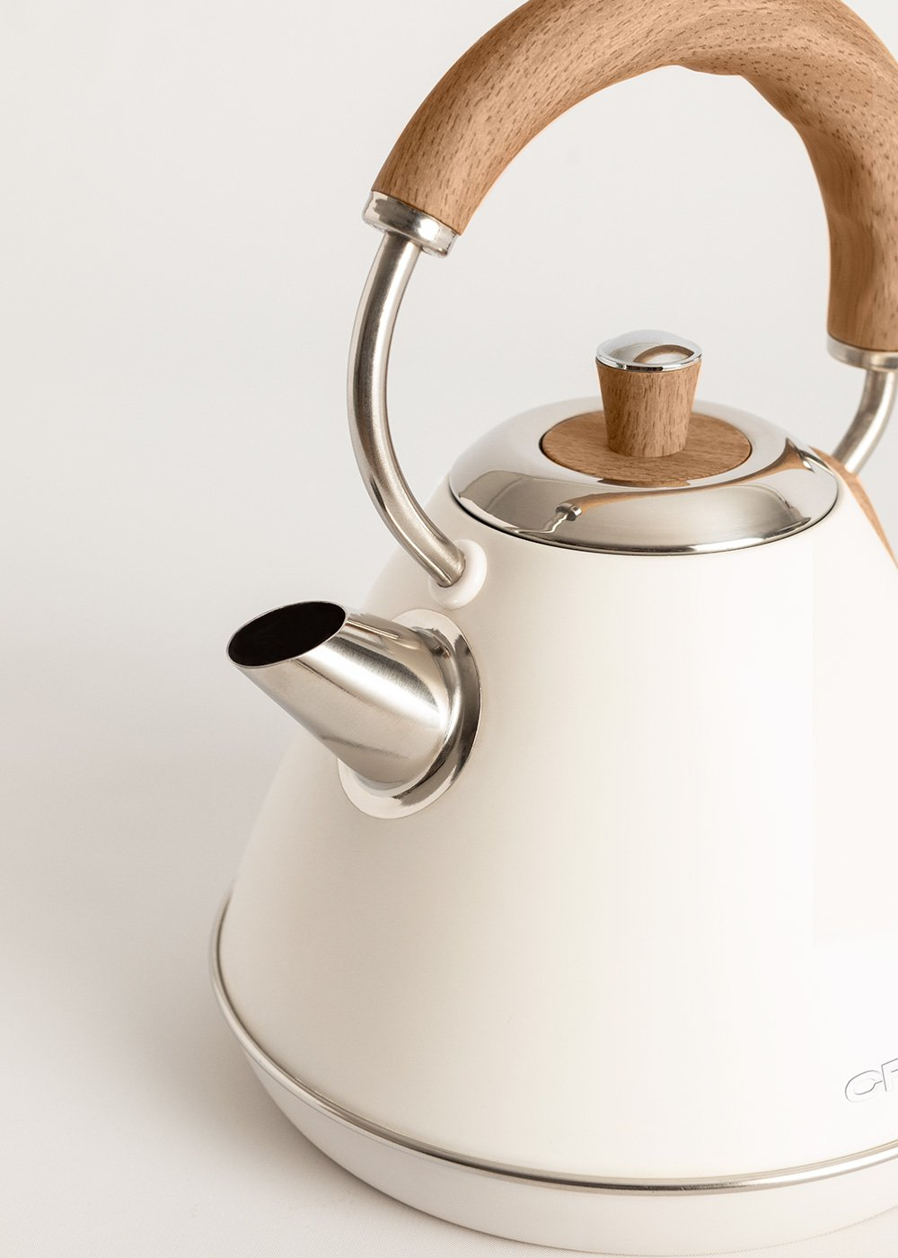 Una tetera crema con asa y tapa de madera. Tiene un pico y base metálicos. El fondo es blanco.