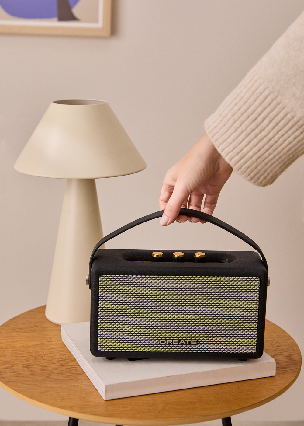 Una mano sostiene un altavoz negro con perillas doradas sobre una mesa de madera. Una lámpara y libros también están sobre la mesa.