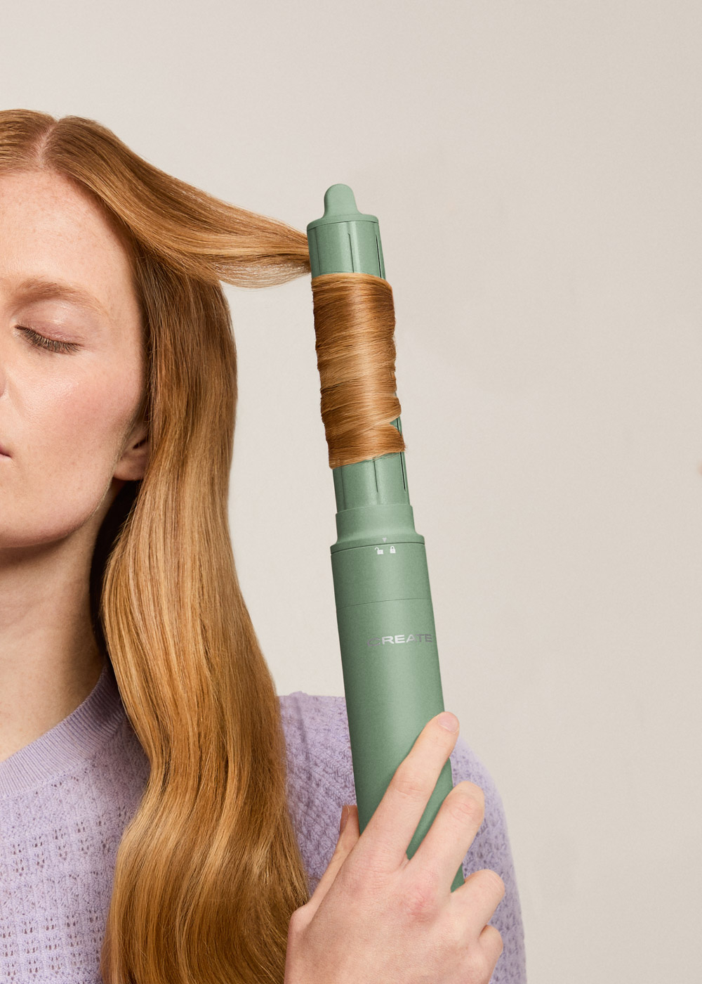 Una mujer se riza el pelo con un rizador verde. Su pelo es largo y rubio. Lleva un suéter morado.