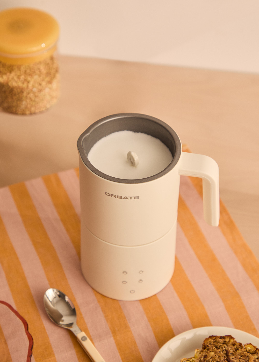 Un espumador de leche crema con asa sobre tela a rayas. Cerca hay una cuchara y un tarro con un plato de pan.
