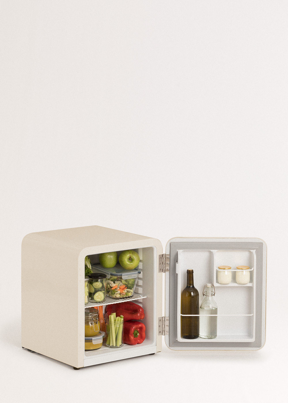 Un pequeño refrigerador crema abierto muestra manzanas, verduras, frascos, botellas y recipientes. El fondo es blanco.