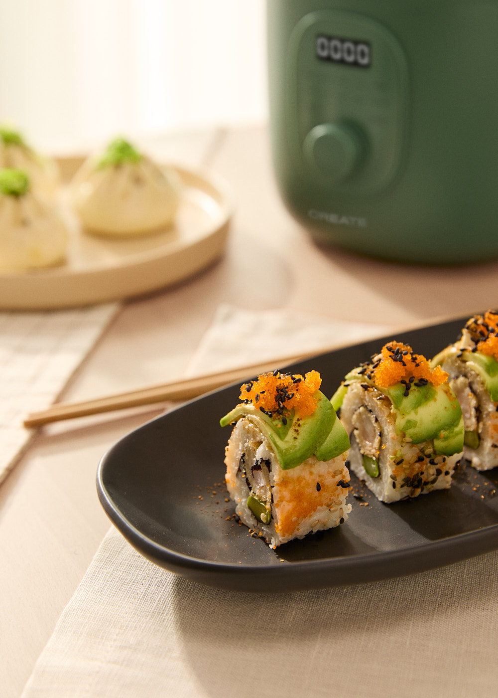 Rollos de sushi con aguacate y huevas naranjas en un plato negro. Dim sum y un electrodoméstico verde en el fondo.