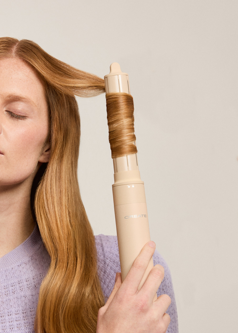 Una mujer con cabello rojo se riza el cabello con un rizador beige. Sus ojos están cerrados. El fondo es de color beige claro.