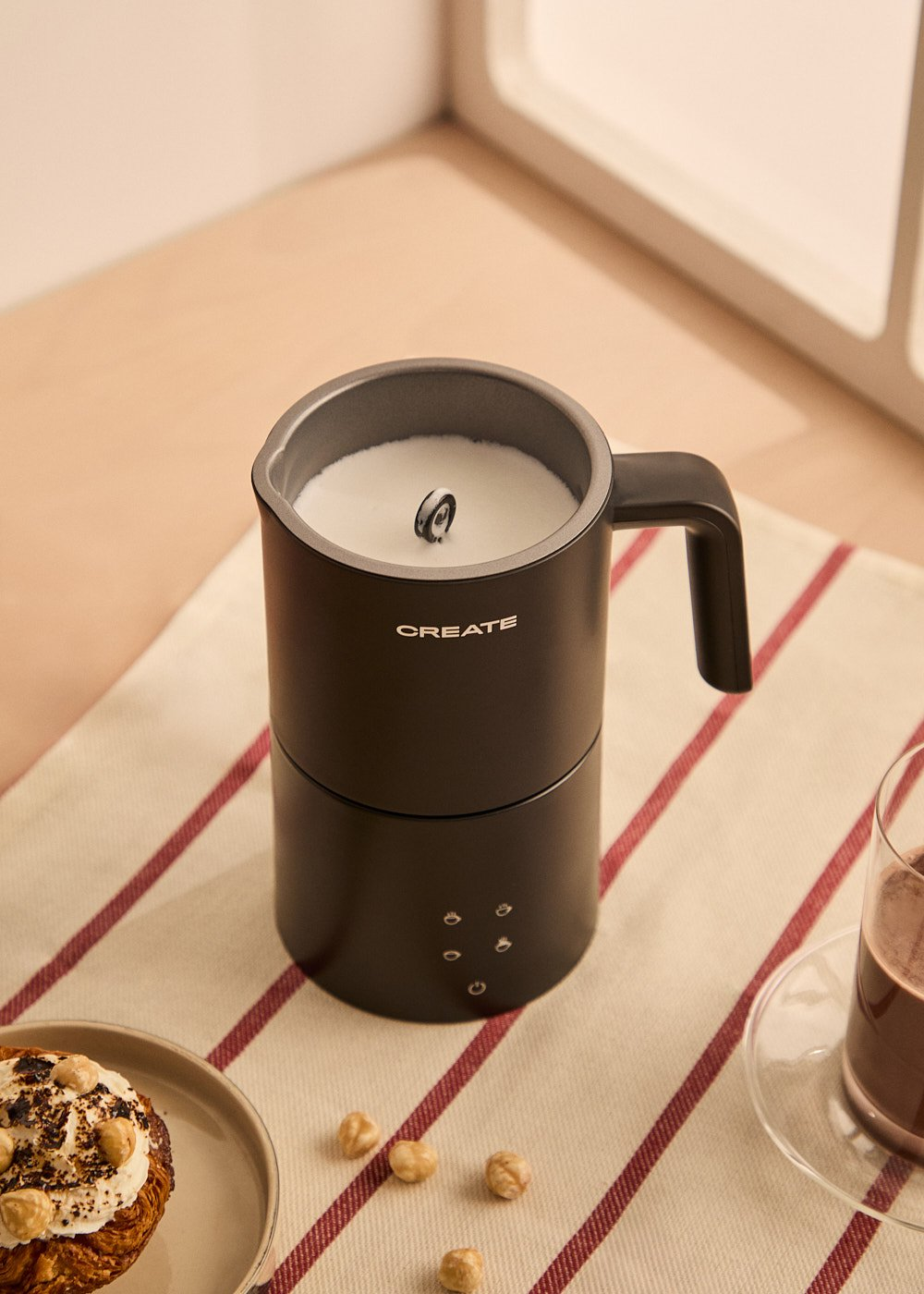 Un espumador de leche negro con asa, un pastel, avellanas y un vaso de chocolate caliente están sobre un mantel a rayas.