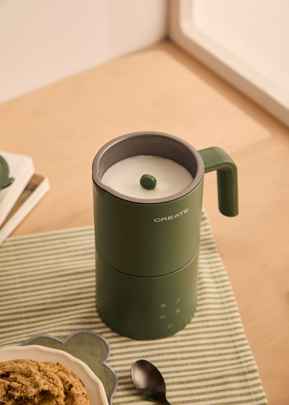 Un espumador de leche verde con leche dentro, y una galleta en un plato con una cuchara. Una servilleta a rayas está debajo del espumador.