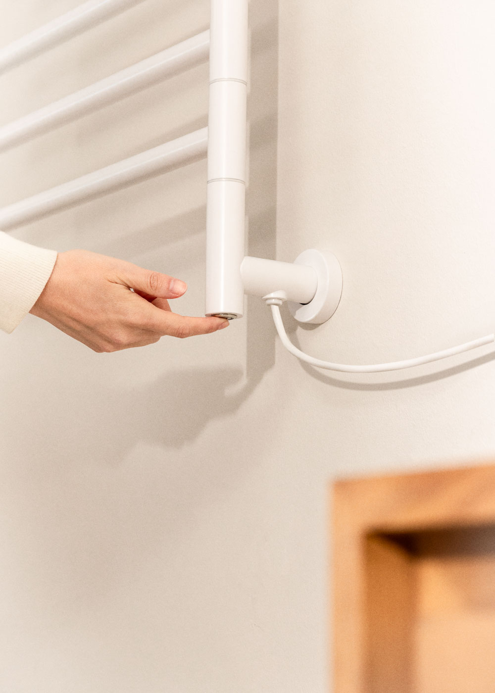 Una mano toca un calentador de toallas blanco en una pared clara. El calentador tiene barras horizontales y un tubo vertical.