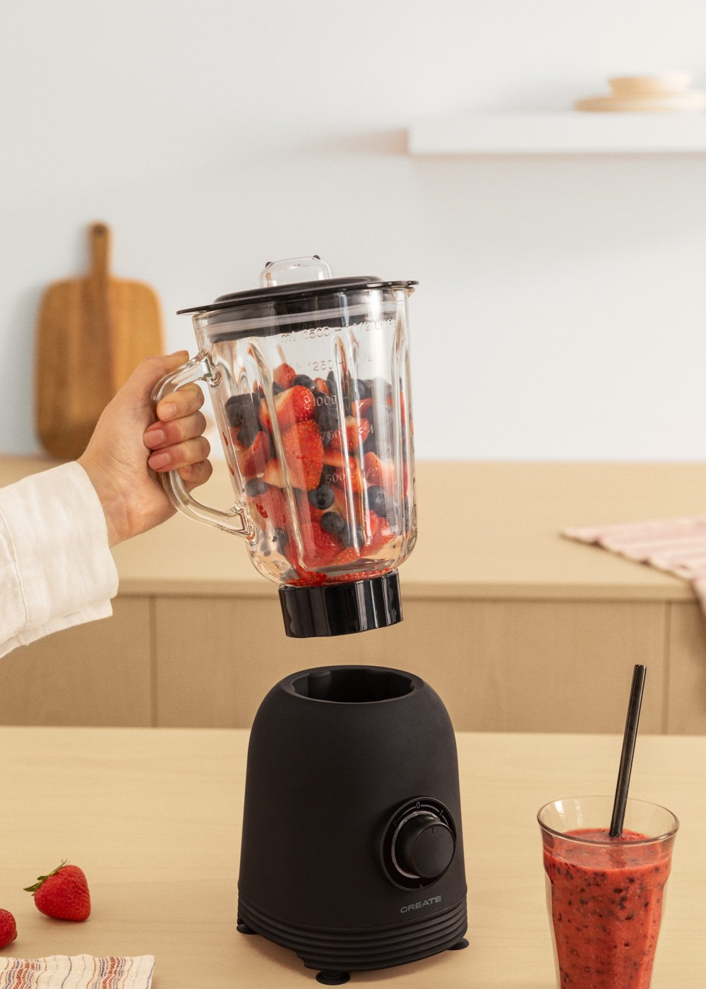 Eine Hand hält einen Mixer, gefüllt mit Beeren. Eine schwarze Mixerbasis befindet sich darunter. Ein Smoothie und Erdbeeren liegen auf dem Tisch.
