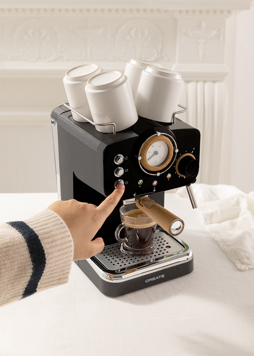 Una mano presiona el botón de una máquina de espresso negra. El café está en una taza de vidrio. Hay tazas blancas encima de la máquina.