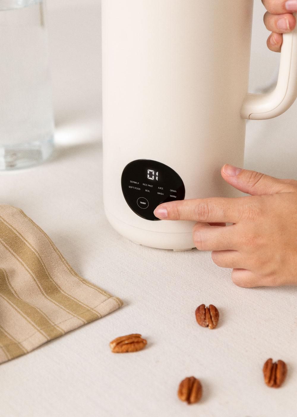 Una mano presiona un botón en un electrodoméstico color crema. Nueces pecanas dispersas sobre la superficie blanca. Un vaso y una servilleta son visibles.
