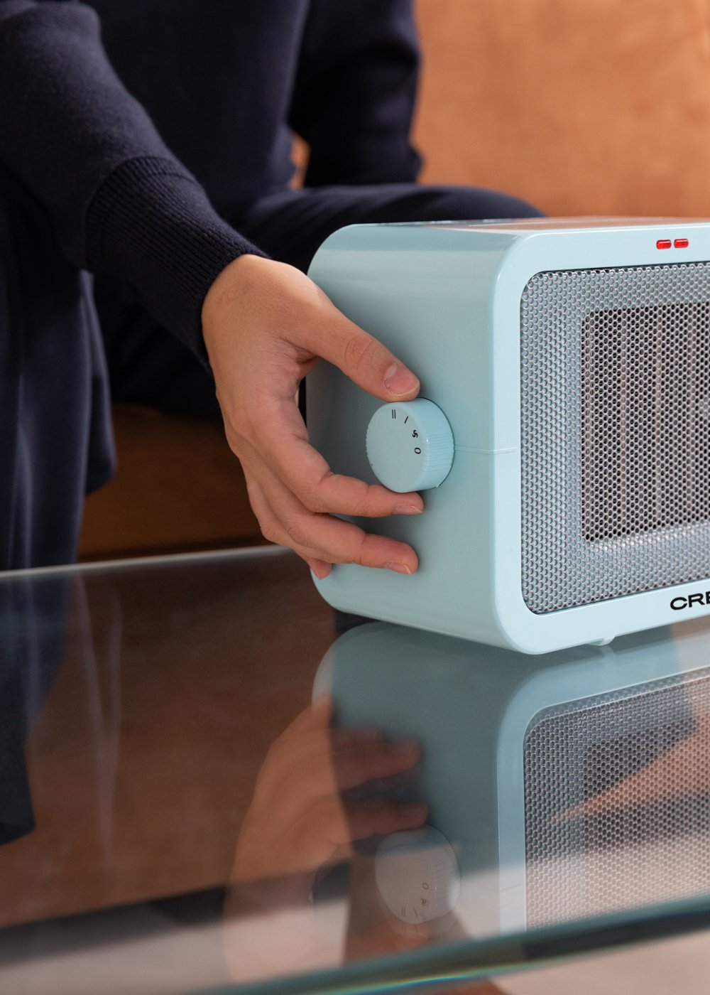 Una persona ajusta un calefactor azul claro sobre una mesa de vidrio. Se gira el dial del calefactor. La escena es interior.