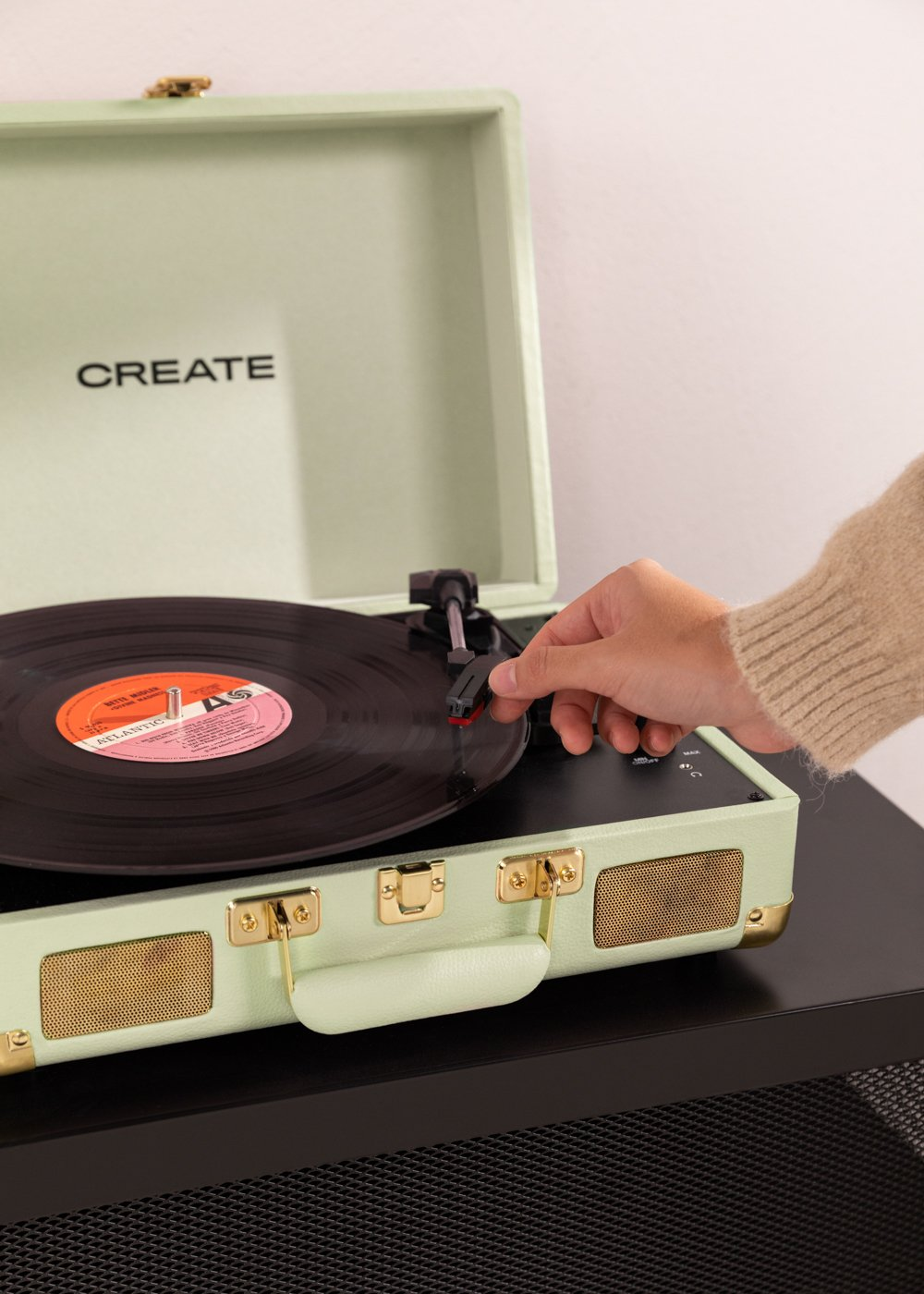 Una mano coloca la aguja en un disco de vinilo en un tocadiscos verde menta. La palabra CREATE es visible.