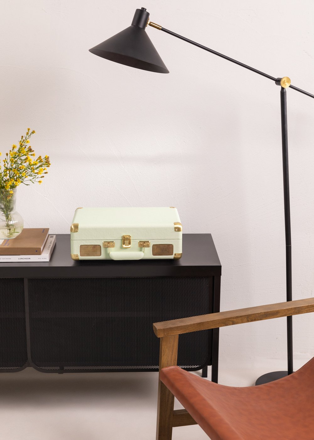 Un tocadiscos verde menta está sobre una mesa negra con flores amarillas y una lámpara. Una silla de madera está a la derecha.