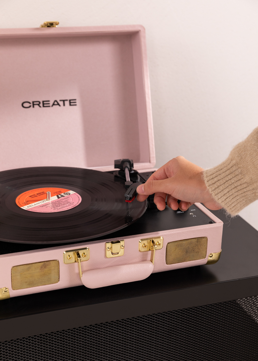 Una mano coloca la aguja en un disco en un tocadiscos rosa. La palabra CREATE es visible. El tocadiscos está sobre una mesa negra.