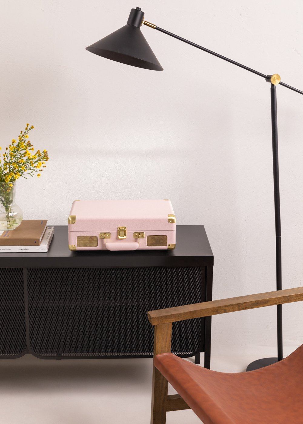 Un tocadiscos rosa sobre una gabinete negro. Una lámpara negra y una silla de madera también son visibles en la habitación.