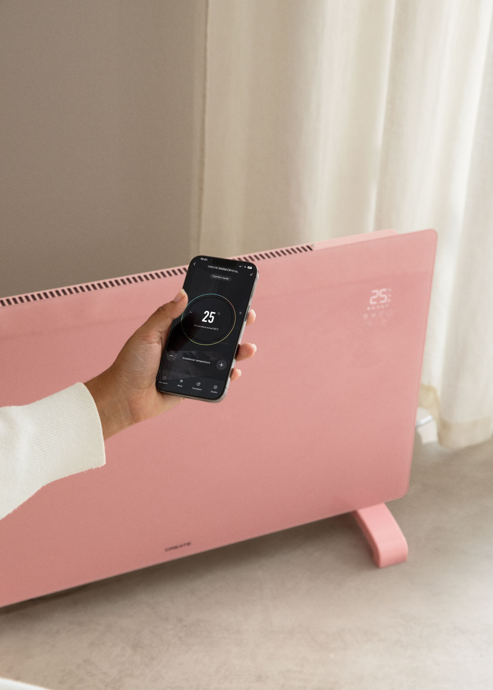 Una mano sostiene un teléfono que muestra 25 grados en un calentador rosa. Manga de suéter blanco visible. Pared beige y cortina blanca.