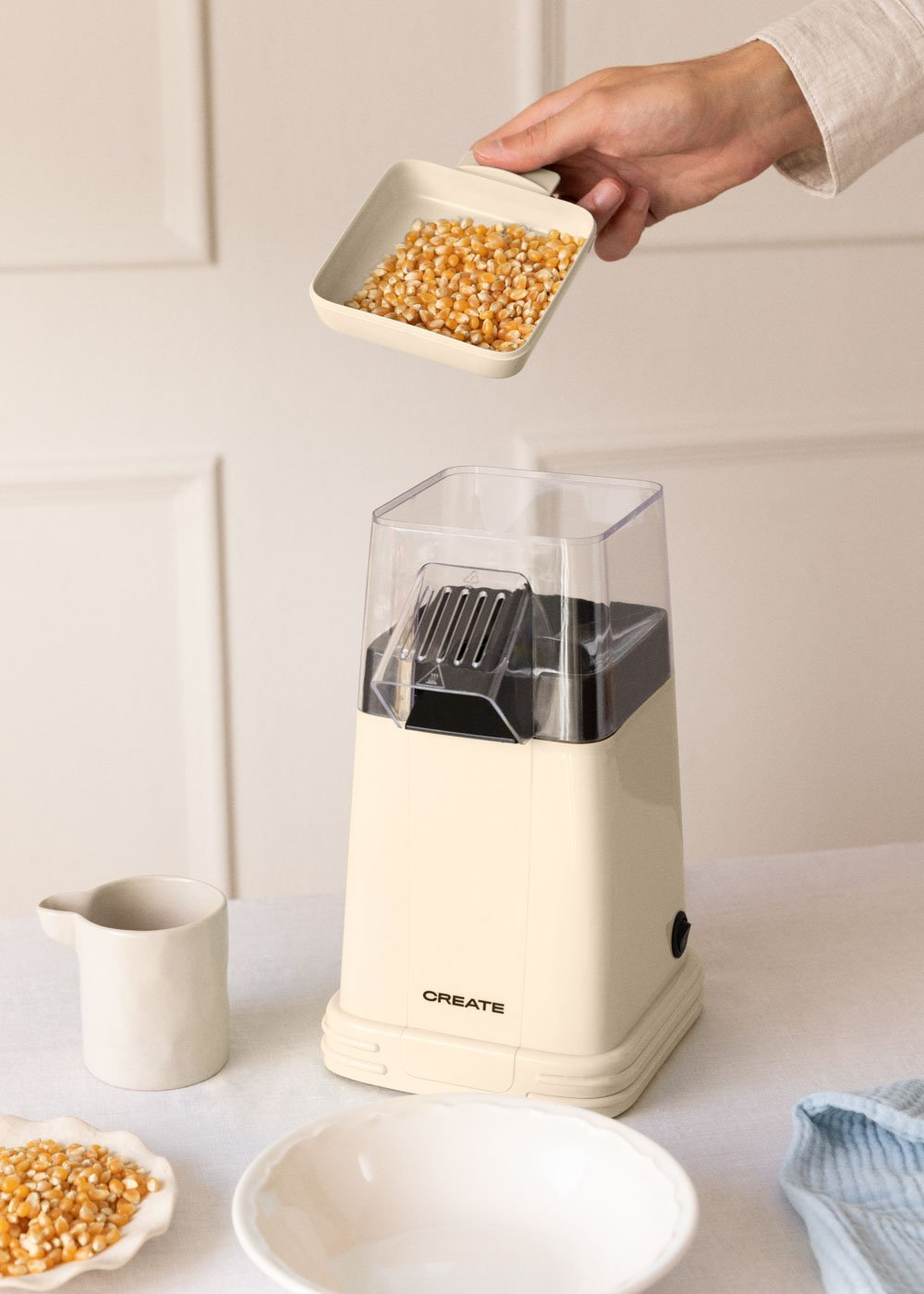 Una mano vierte granos de maíz en una máquina de palomitas de color crema. Otros granos y cuencos están en el marco, contra un fondo blanco.