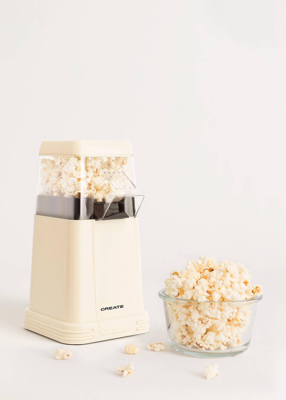 Una máquina de palomitas crema y un cuenco de cristal lleno de palomitas sobre una superficie blanca. Algunas palomitas esparcidas.