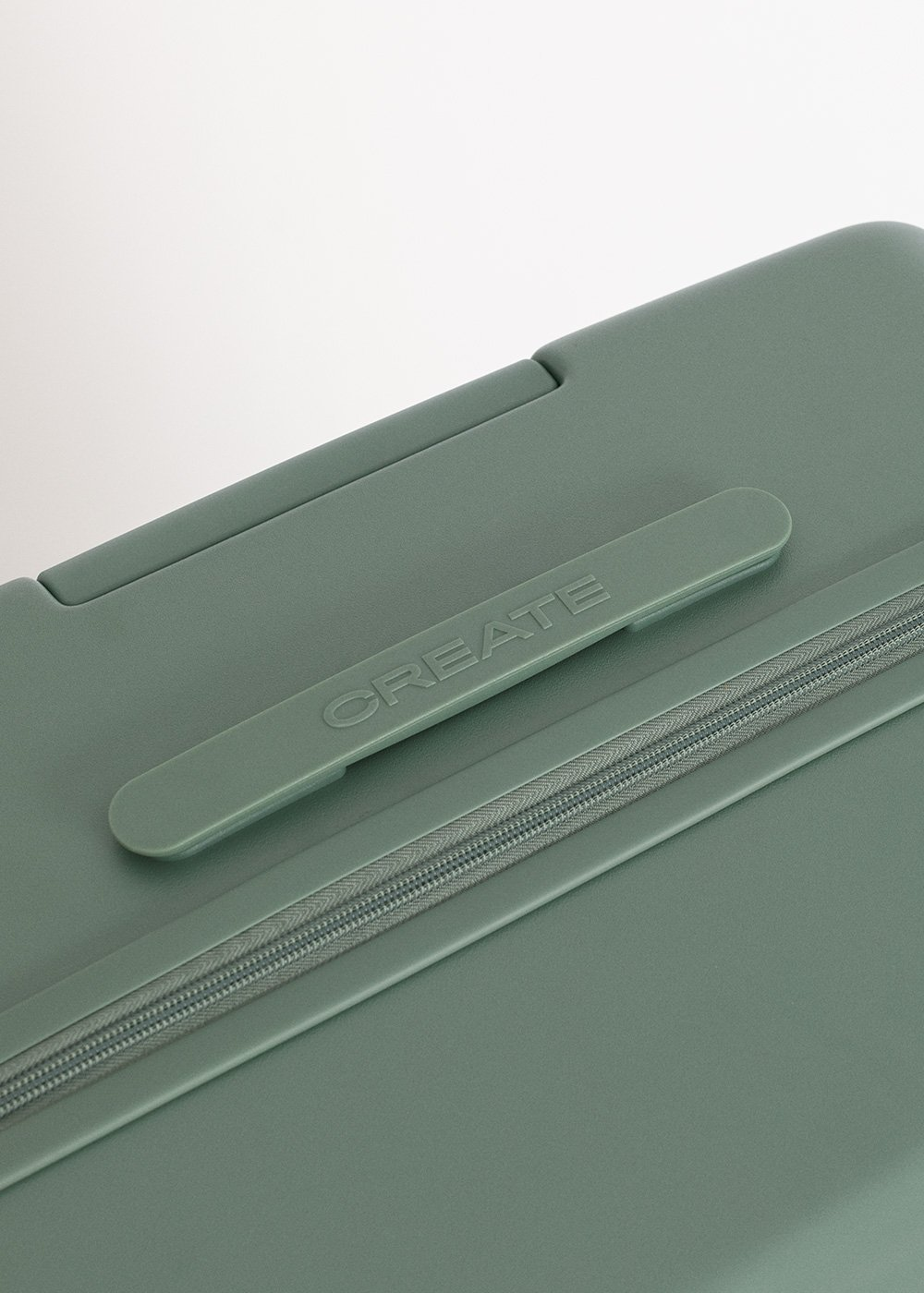 Primer plano de una maleta verde con la palabra CREATE. La cremallera es visible. El fondo es blanco.