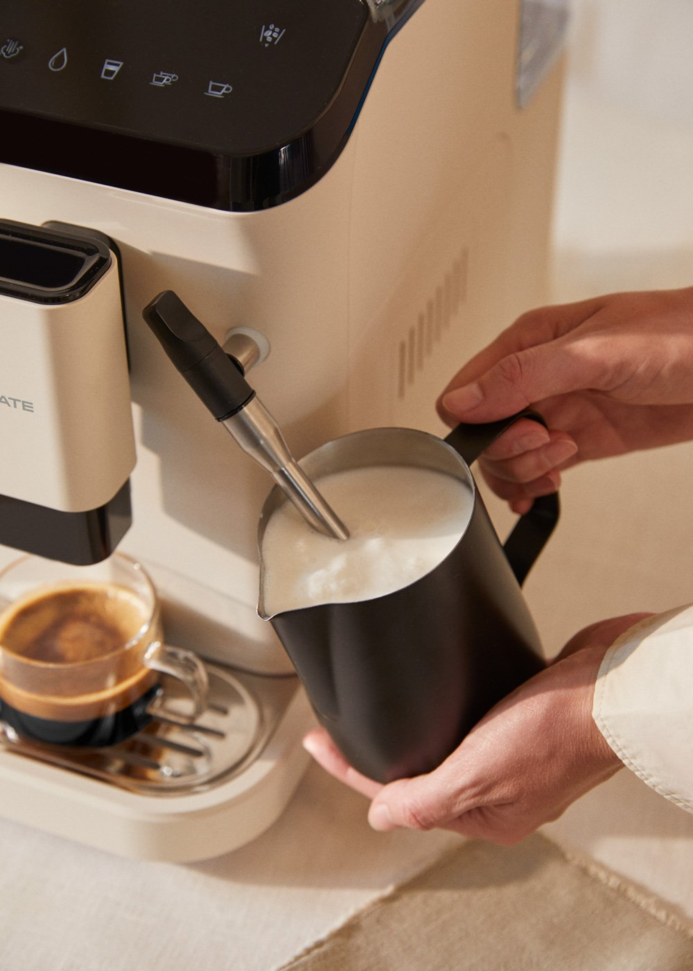 Una persona vierte leche espumosa de una jarra negra en un vaso transparente. También se ven una cafetera espresso y una taza de café.