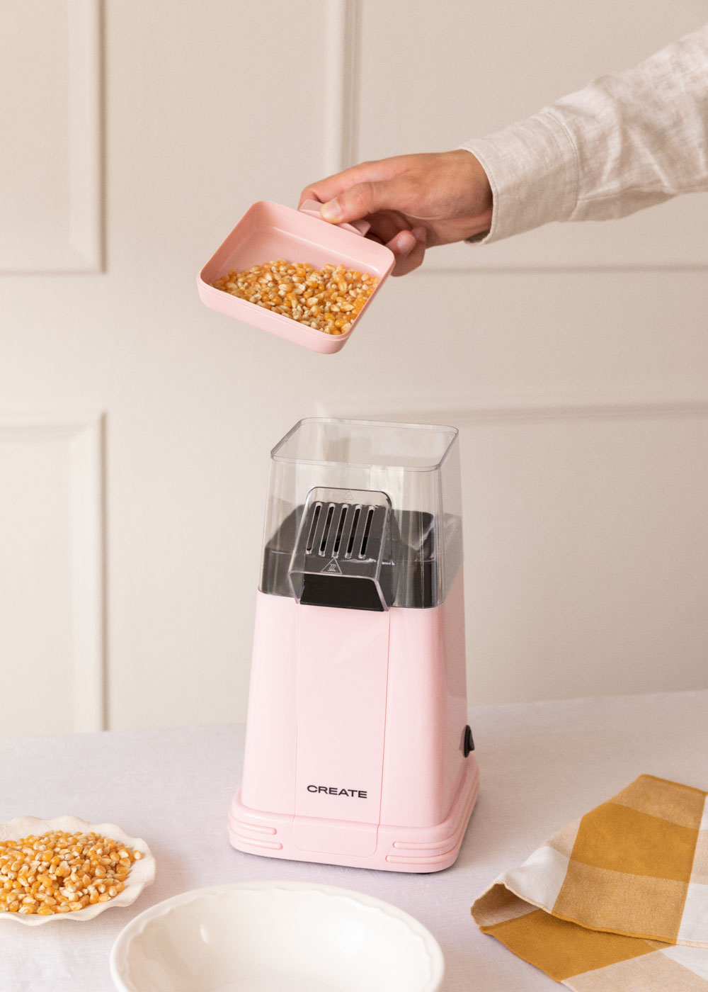 Una mano vierte granos de maíz en una máquina de palomitas de maíz rosa. También hay un cuenco con granos y una servilleta a cuadros.