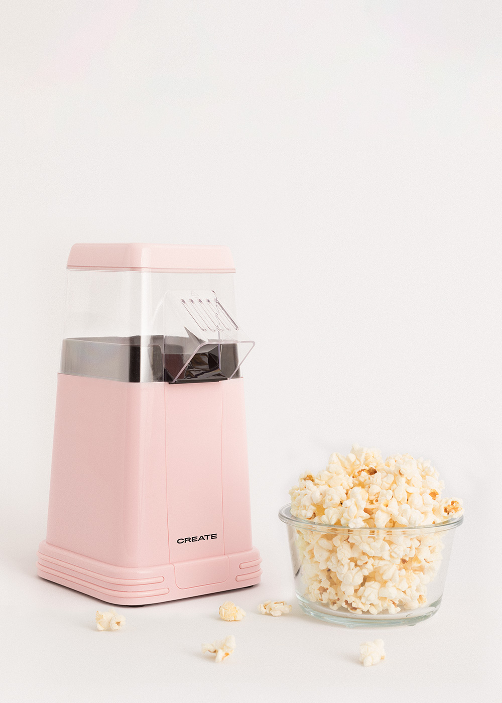 Una máquina de palomitas rosa y un cuenco de cristal con palomitas sobre una superficie blanca. Algunas palomitas esparcidas.