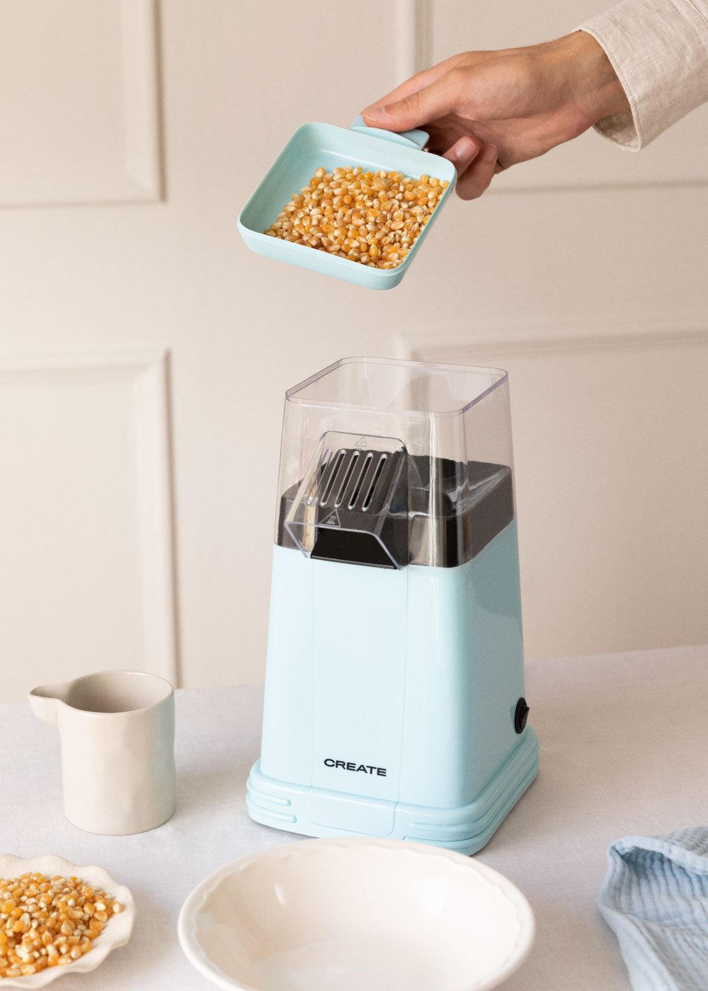 Una mano vierte granos de maíz en una máquina de palomitas de maíz azul claro. Otros elementos incluyen una taza, un cuenco y un plato con maíz.
