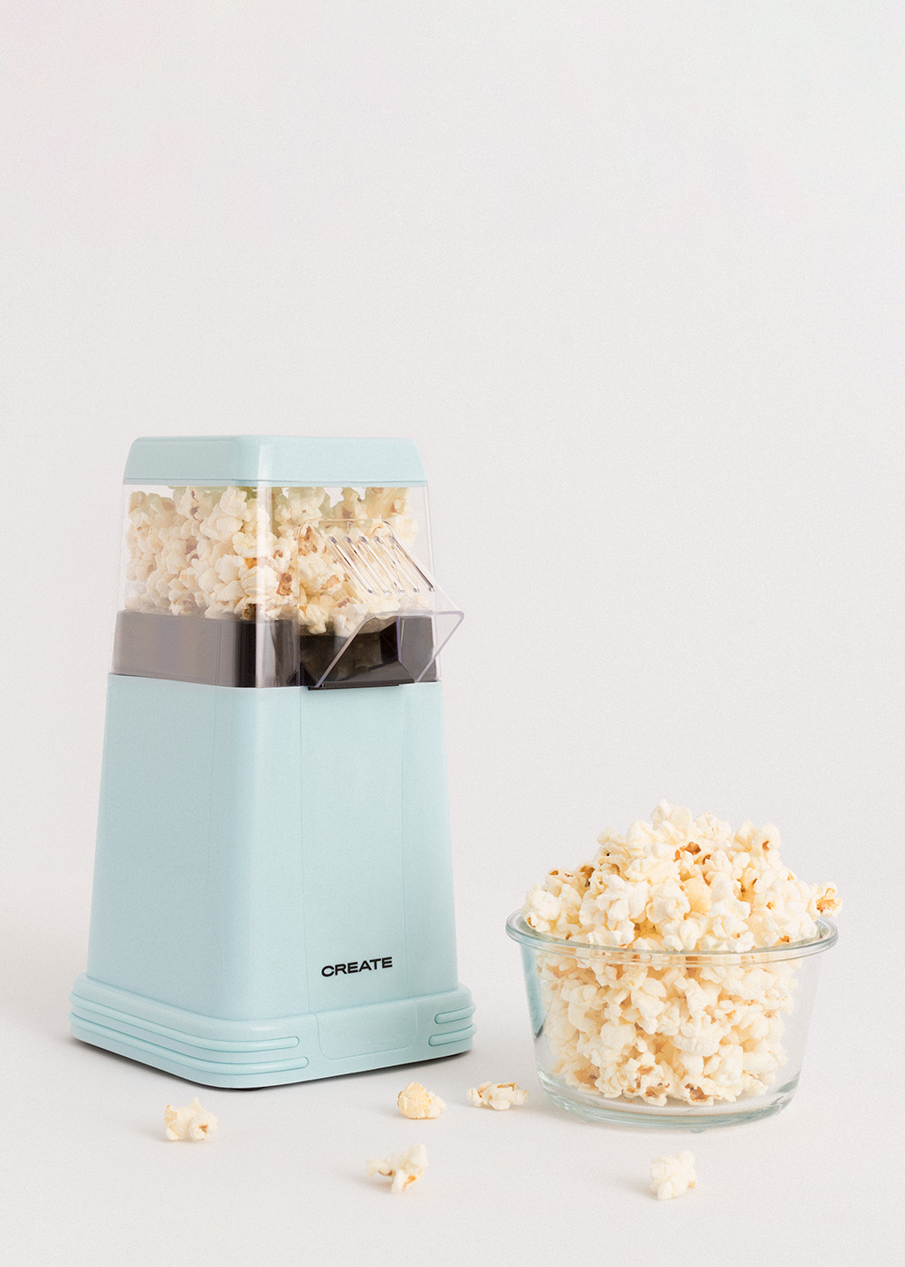 Una máquina de palomitas de maíz azul claro y un cuenco de vidrio lleno de palomitas sobre una superficie blanca. Algunas palomitas están esparcidas.