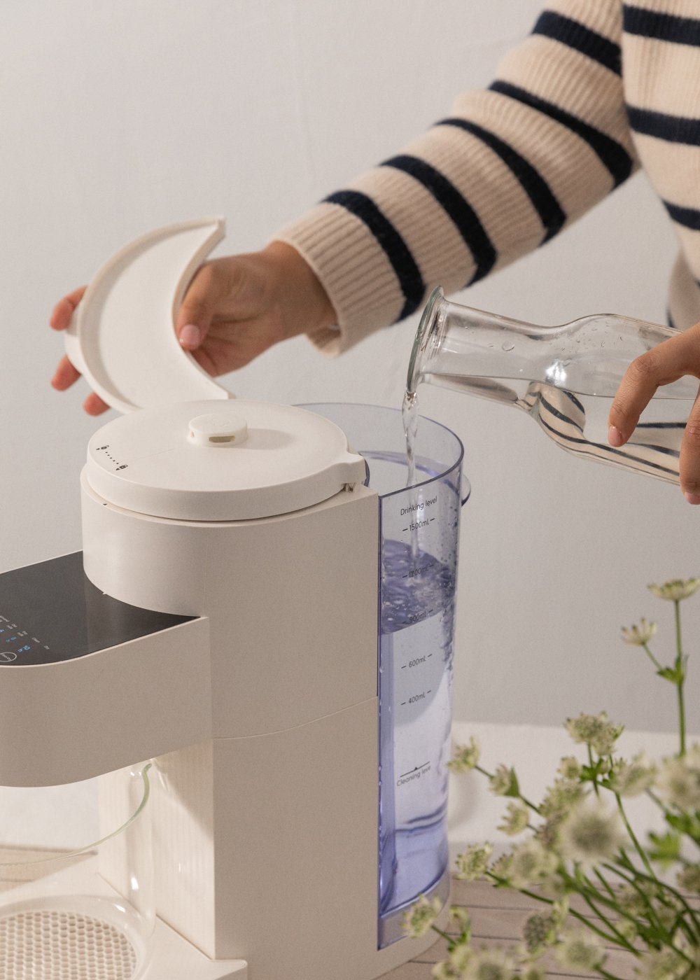 Una persona vierte agua de una jarra de vidrio en un purificador de agua blanco. La persona lleva un suéter a rayas.