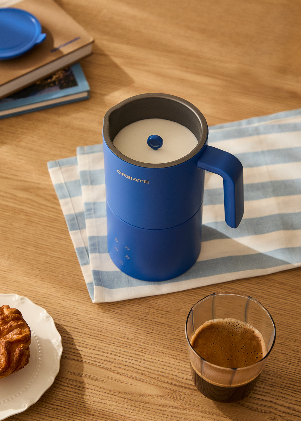 Un espumador de leche azul con asa, una taza de café y un cruasán en un plato sobre una mesa de madera.
