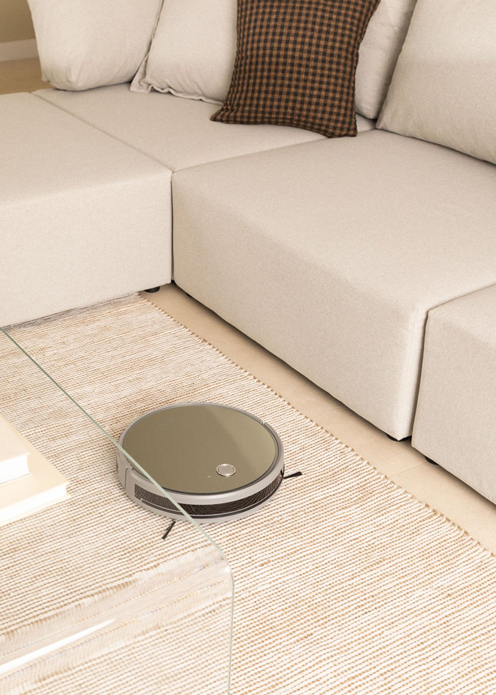 Una aspiradora robot en una alfombra cerca de un sofá seccional beige. También se ven una mesa de vidrio y almohadas.