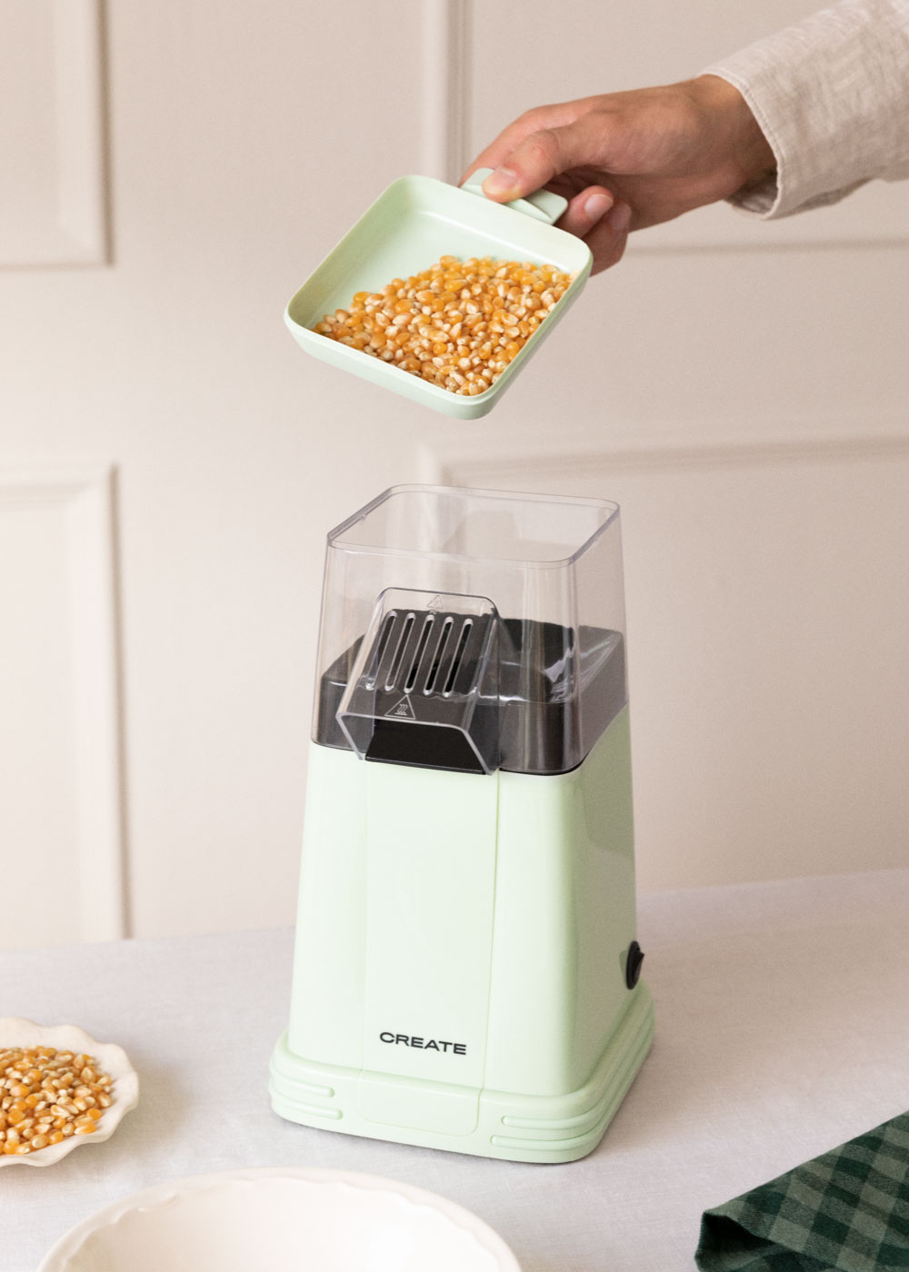 Una mano vierte granos de palomitas de una bandeja verde en una máquina de palomitas verde. Un cuenco blanco y una servilleta verde están en la mesa.