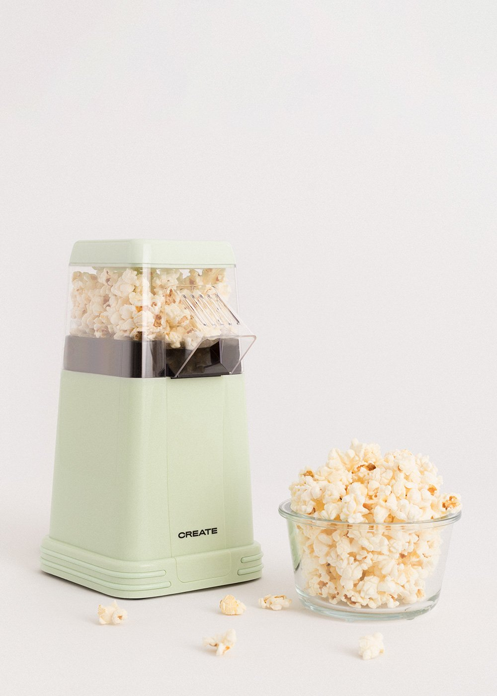 Una máquina de palomitas verde menta con palomitas de maíz dentro y un cuenco de vidrio lleno de palomitas, más granos sueltos.