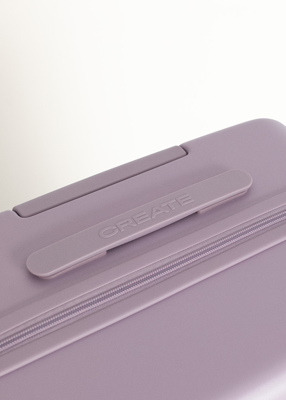 Primer plano de una maleta lavanda, con un asa con 'CREATE' y una cremallera. El fondo es blanco.