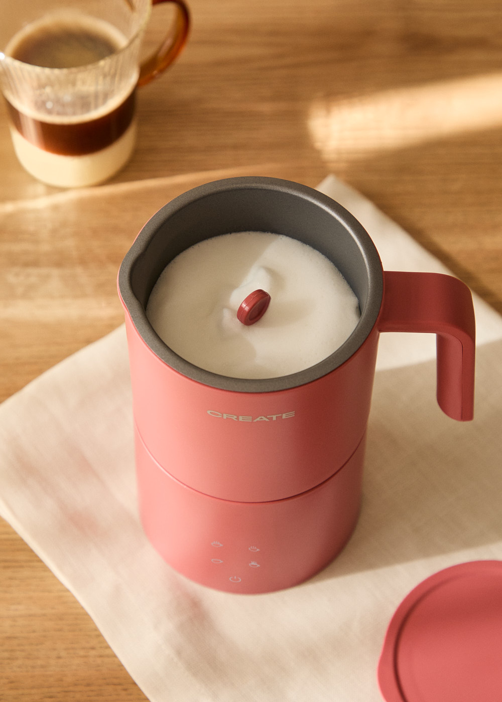 Un espumador de leche rosa con espuma blanca y un botón rojo en la parte superior. Un vaso de café en el fondo sobre una superficie de madera.