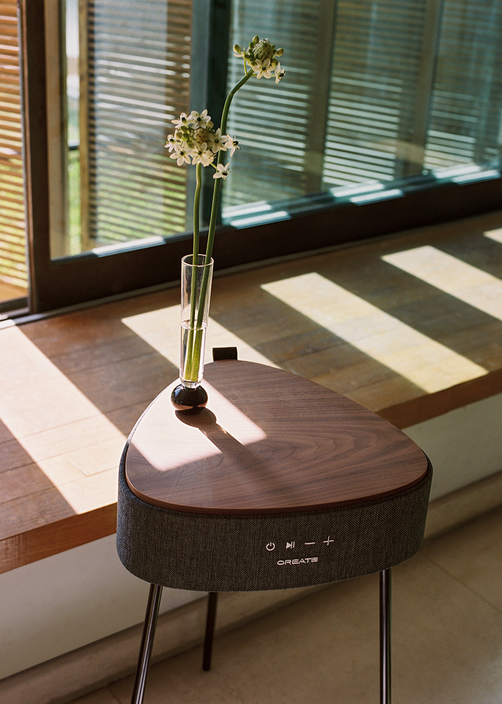 Una mesa moderna con flores, un jarrón de cristal y controles de altavoz. La luz del sol entra por una ventana con persianas en el fondo.