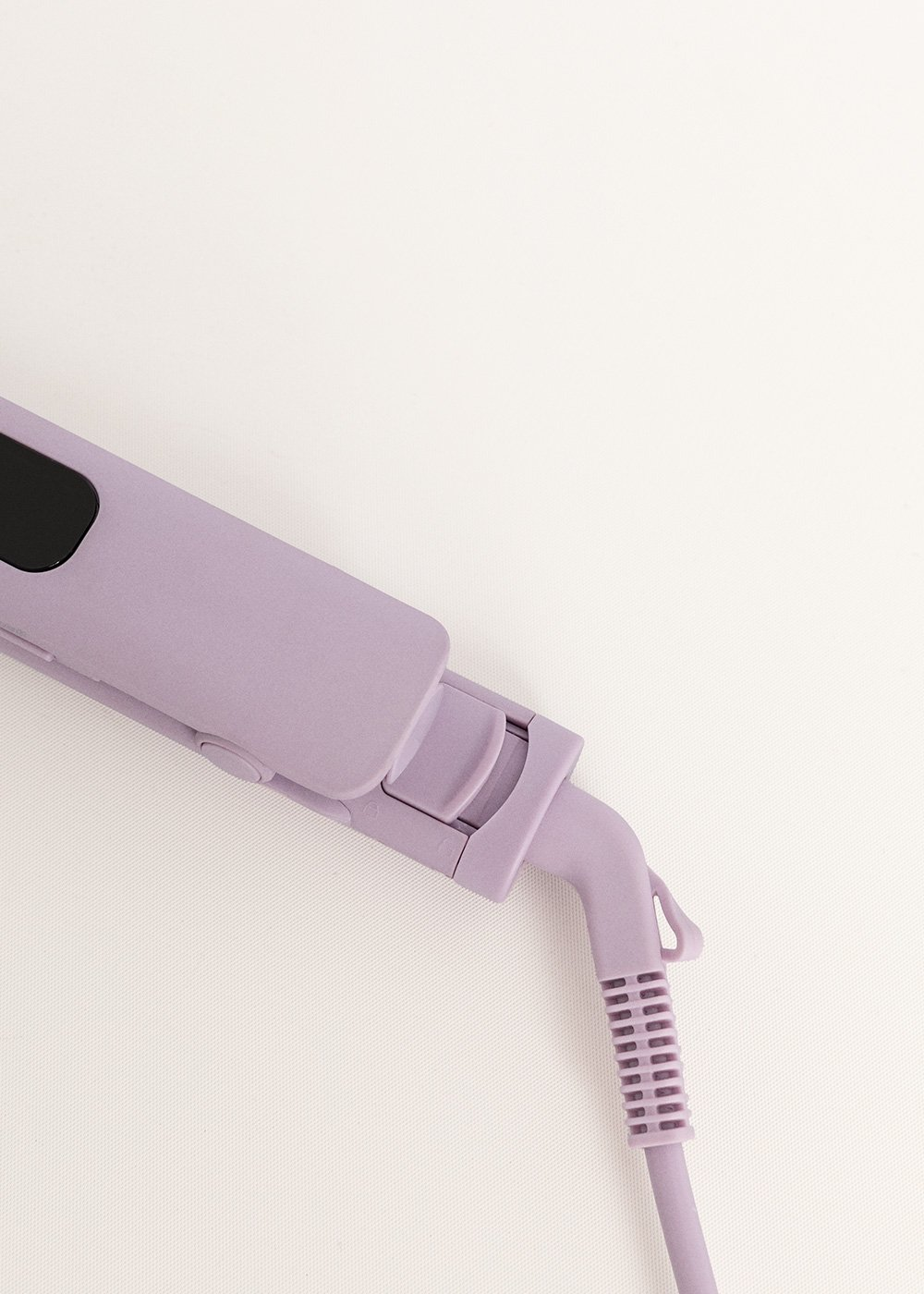Una plancha de pelo lavanda está sobre una superficie blanca. La plancha tiene un panel negro y un cable.