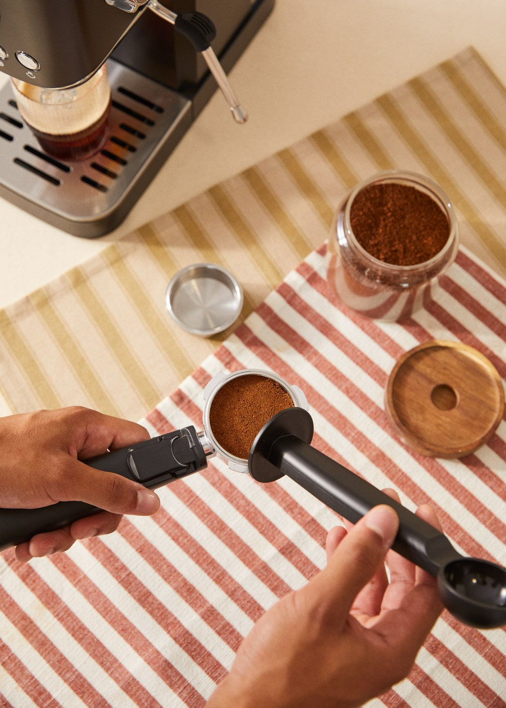 Manos llenando un portafiltro con café molido. Cerca hay una máquina de espresso, un tarro de café y un posavasos de madera.