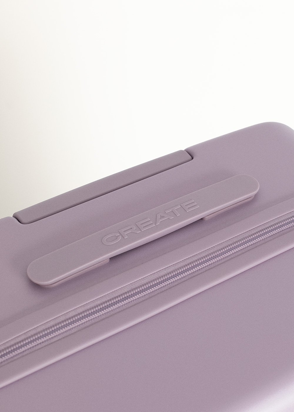 Primer plano de una maleta lavanda. El asa dice 'CREATE'. El detalle de la cremallera es visible. El fondo es blanco.