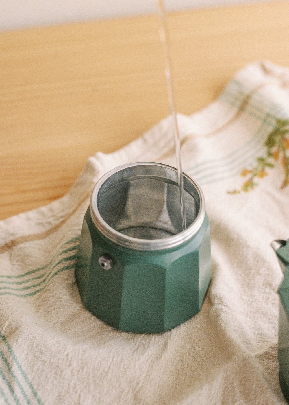Agua vertiéndose en una cafetera moka verde. La olla está sobre un paño blanco con rayas verdes, sobre una superficie de madera.