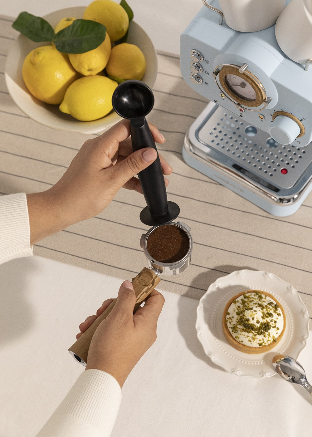 Una persona usa un tamper y portafiltro para preparar café. Se ven un plato de limones, una máquina de espresso azul y un postre.