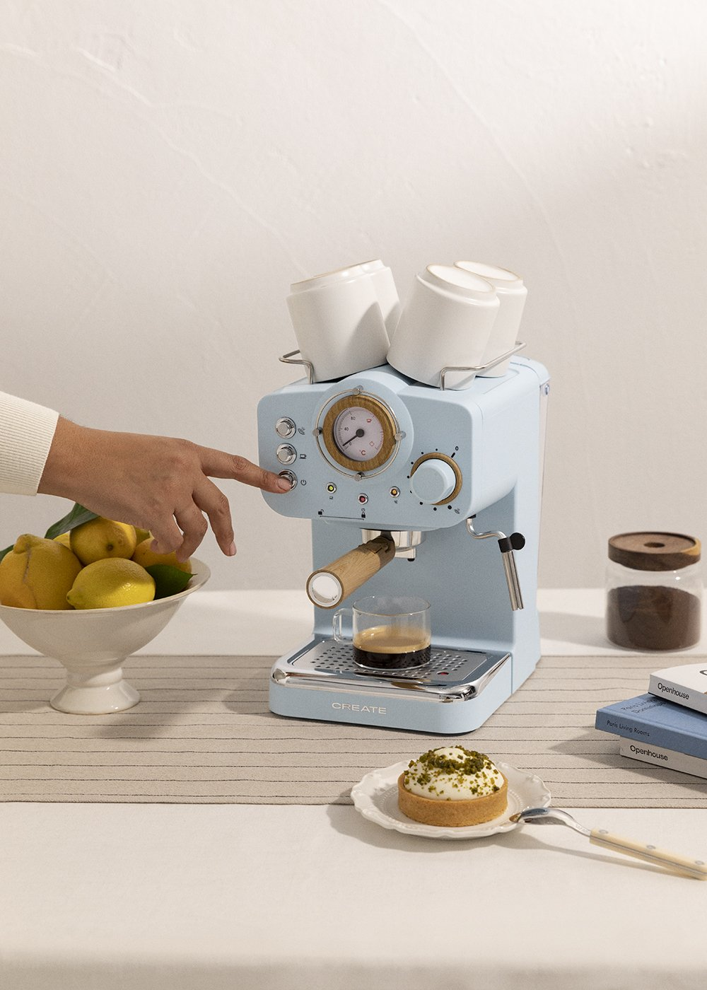 Una persona presionando un botón en una máquina de espresso azul. Una taza de café y una tarta están en la mesa. Limones en un cuenco.