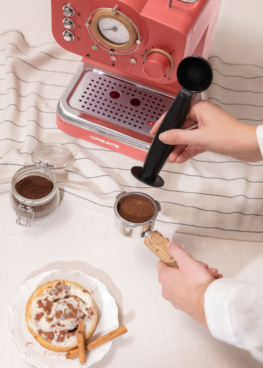 Una persona preparando café con una máquina de espresso. Hay café molido, un pastel y palitos de canela en la mesa.