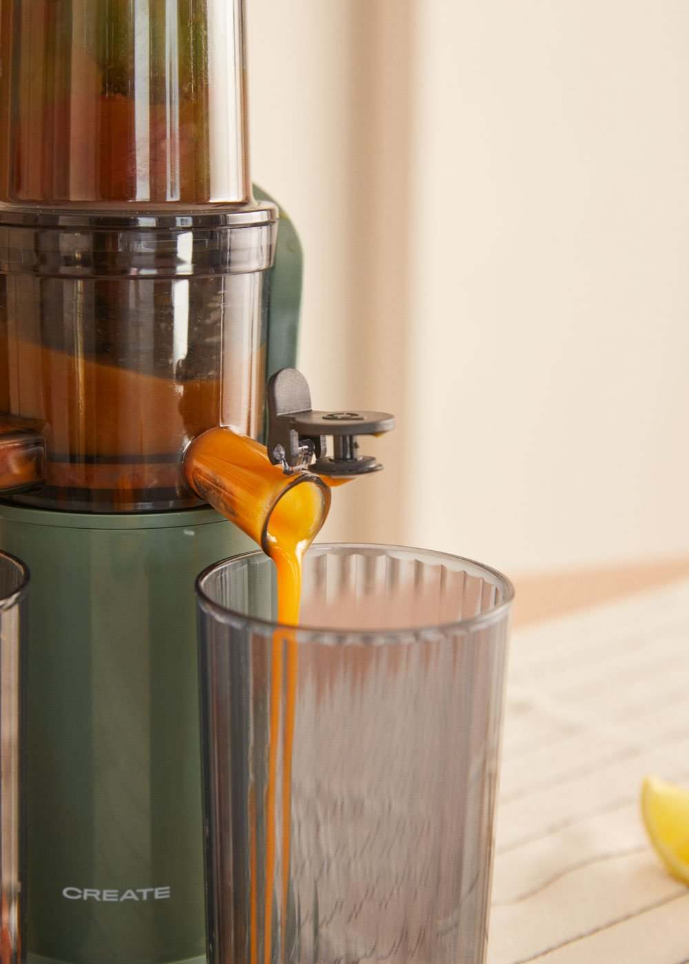 Una licuadora vierte jugo de naranja en un vaso transparente. La licuadora es verde. Una rodaja de limón está sobre una superficie rayada.