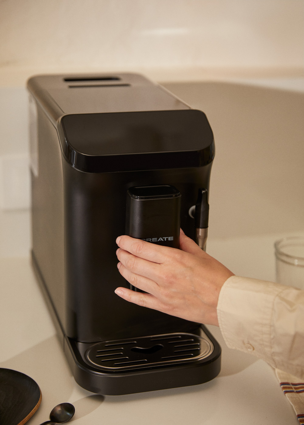 Una mano opera una cafetera negra. La máquina está sobre una encimera de color claro. Se ven una taza y una cuchara.