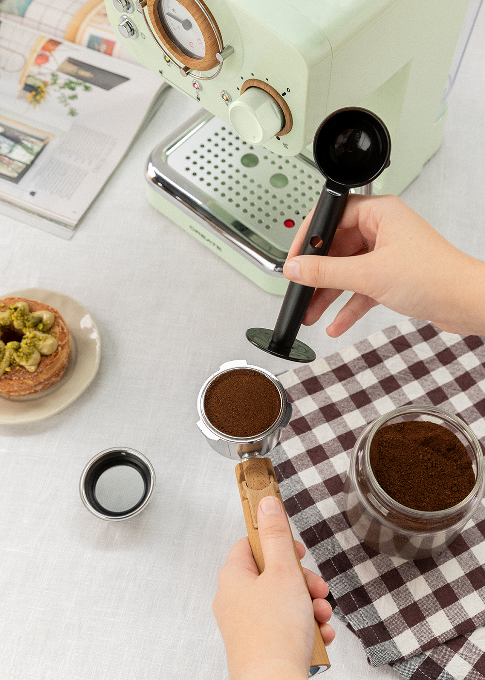 Una persona prepara café con una máquina de espresso. Hay un portafiltro con café molido, una cafetera y un tarro de cristal.