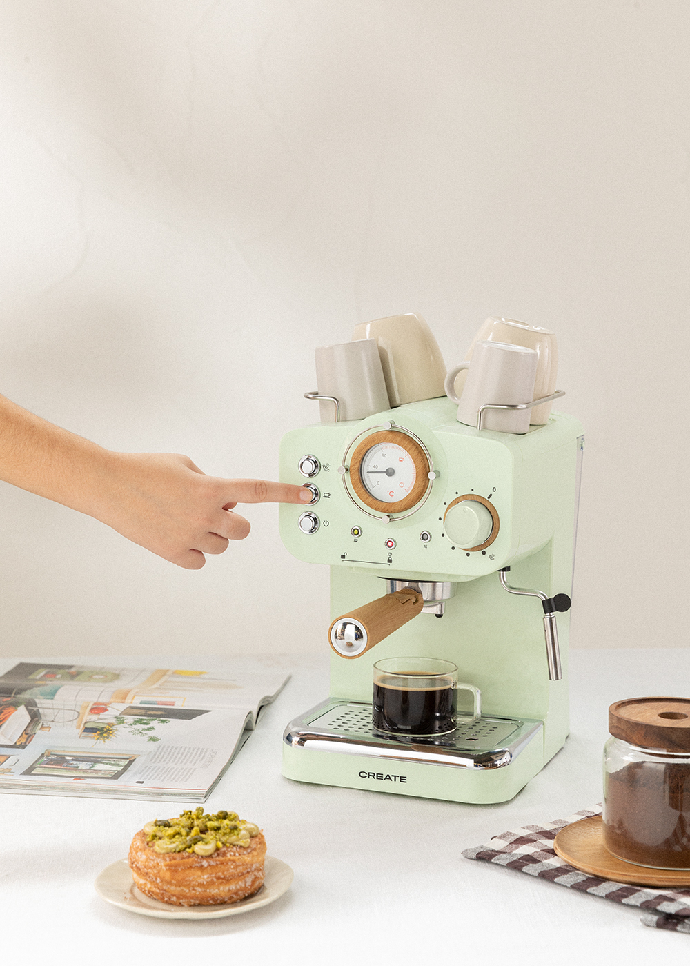 Una mano presiona un botón en una máquina de espresso verde menta. Un vaso de café y un pastel están sobre la mesa.