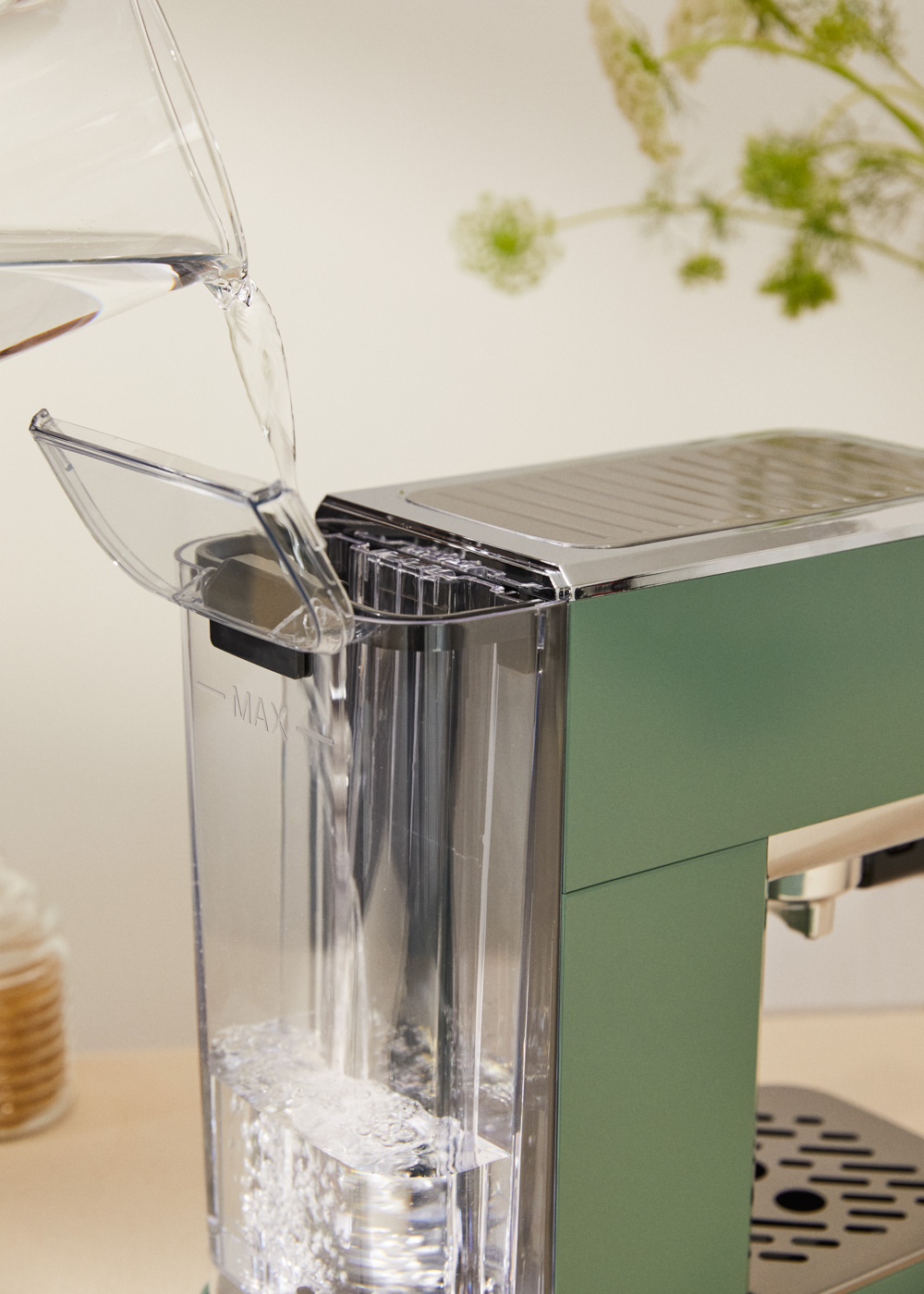 Agua vertiéndose en el depósito de agua de una máquina de espresso verde. Tanque de plástico transparente y parte superior plateada. Fondo floral.