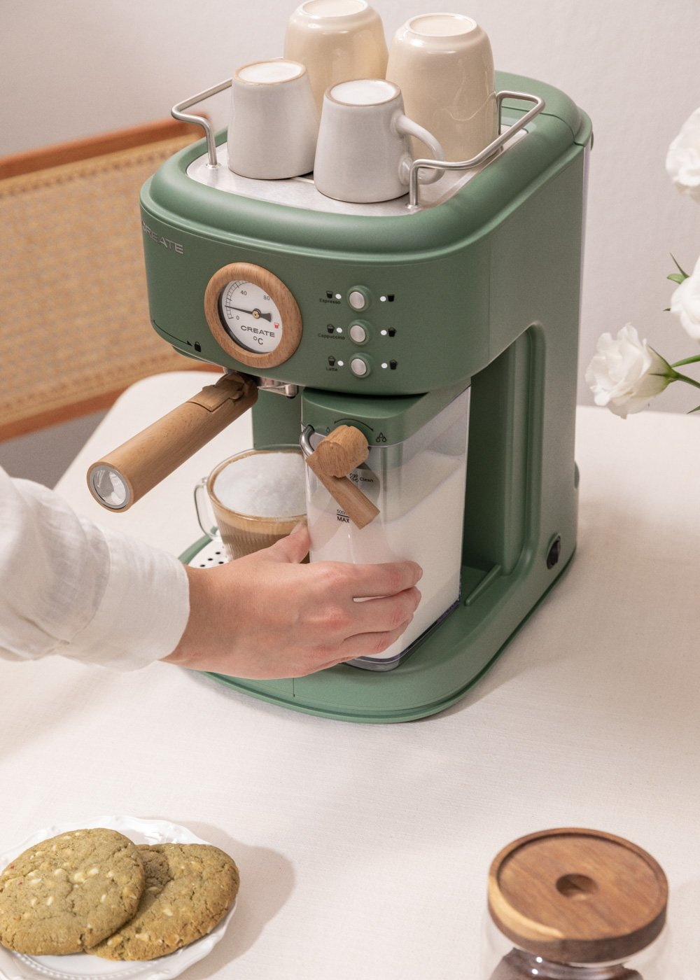 Cafetera verde con tazas, mano alcanzando un recipiente. Galletas y flores sobre superficie blanca.